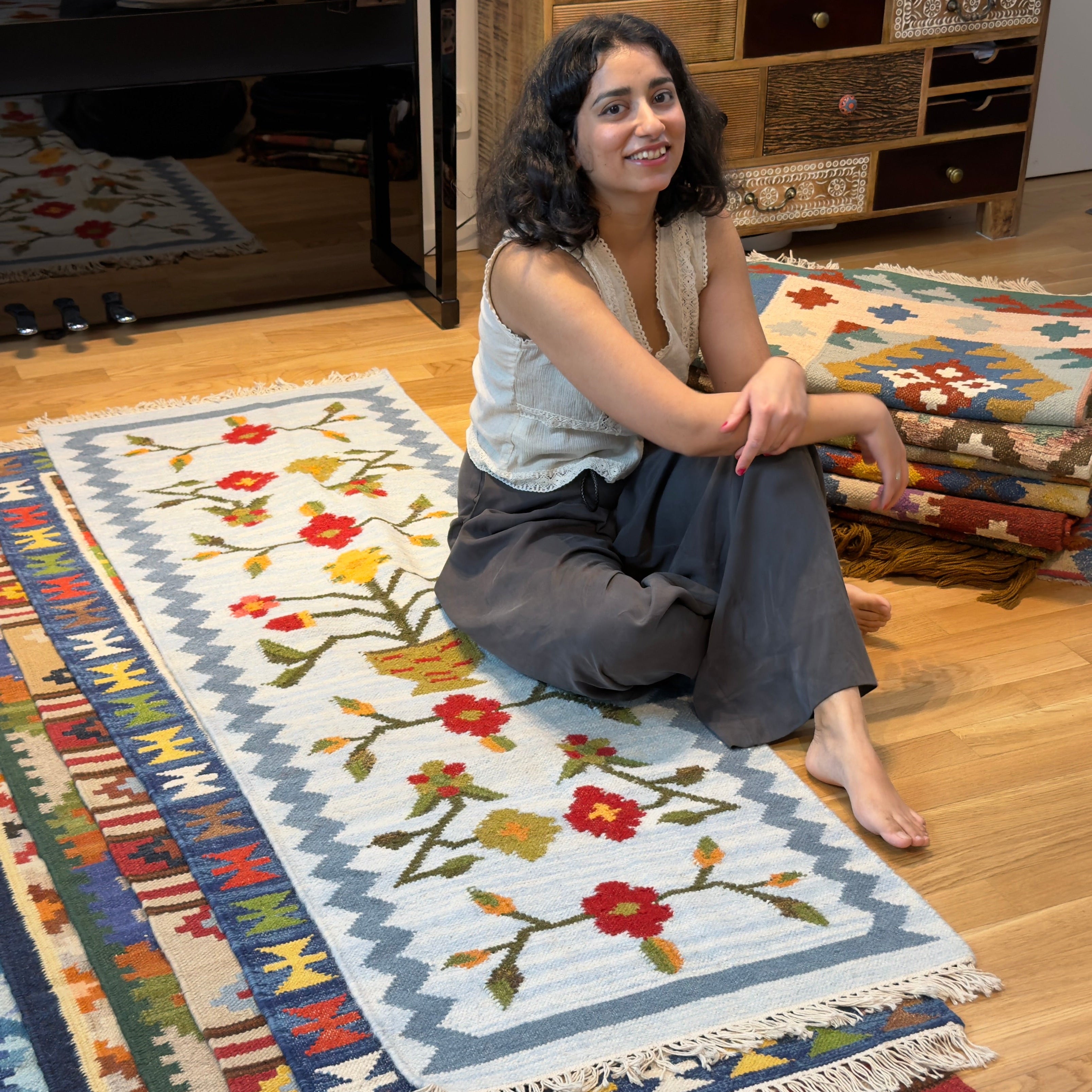 A woman sits barefoot on a wooden floor in a home interior, surrounded by several colorful patterned rugs. She is wearing a white sleeveless top and dark gray pants.
